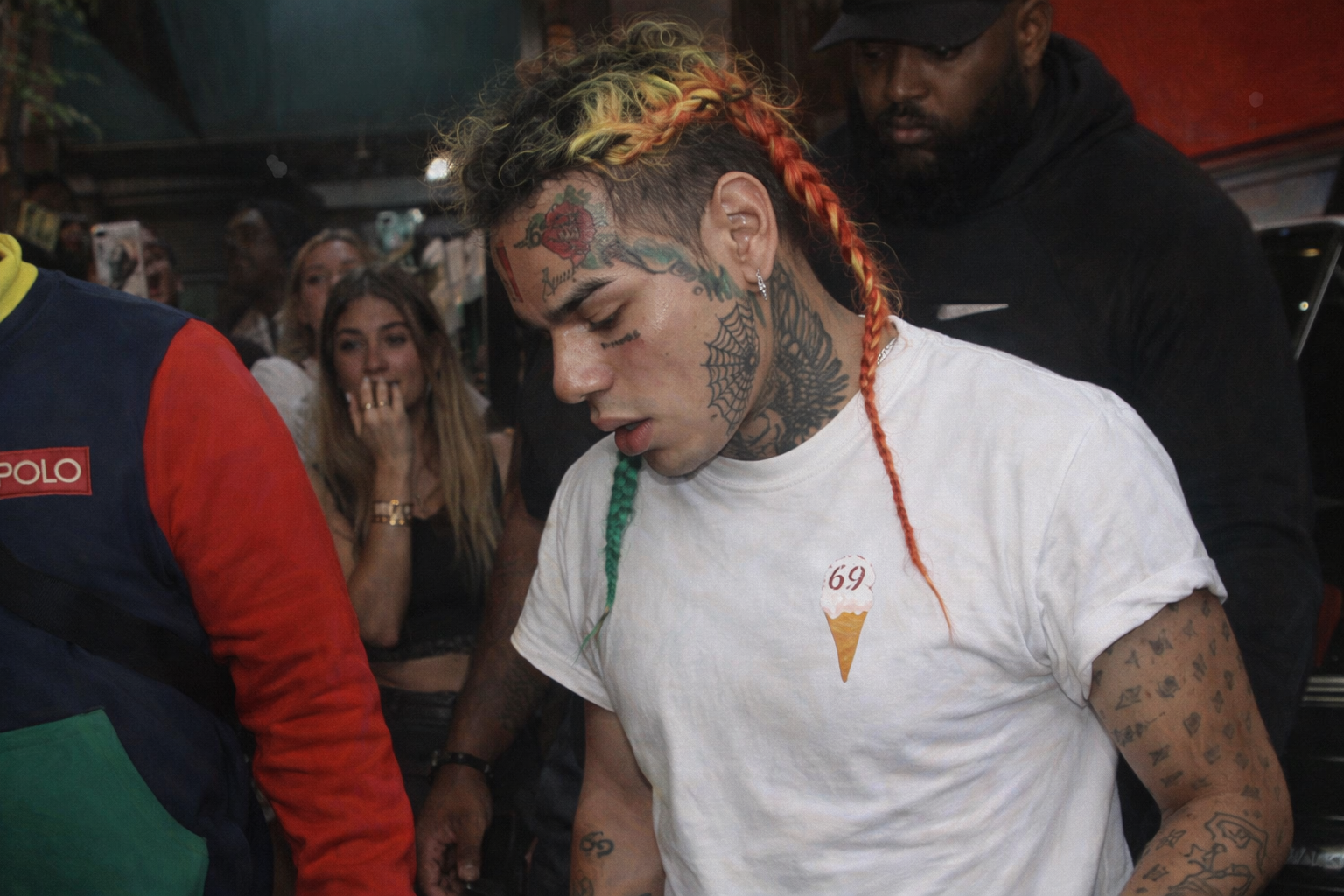 6ix9ine 現身人群中,彩色編髮與臉部刺青明顯,身穿白色 T 恤,低頭行走於公共場合