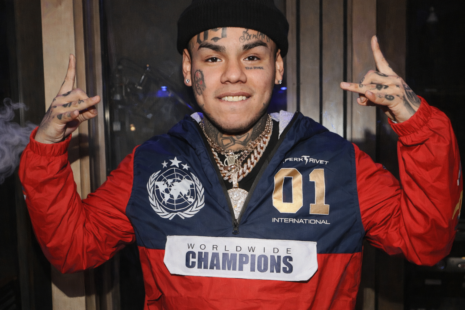 6ix9ine 於室內場合露面,配戴黑色毛帽與多條項鍊,身穿紅藍拼色外套,雙手比出手勢